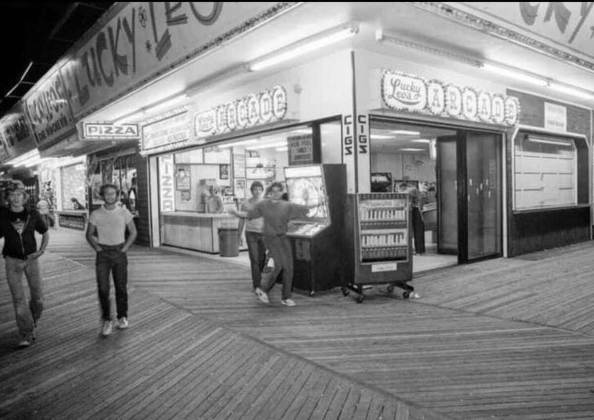 A black-and-white photo of an old-fashioned boardwalk arcade with a lighted sign, a cigarette vending machine outside, and a few people standing near the entrance and walking along the wooden planks.