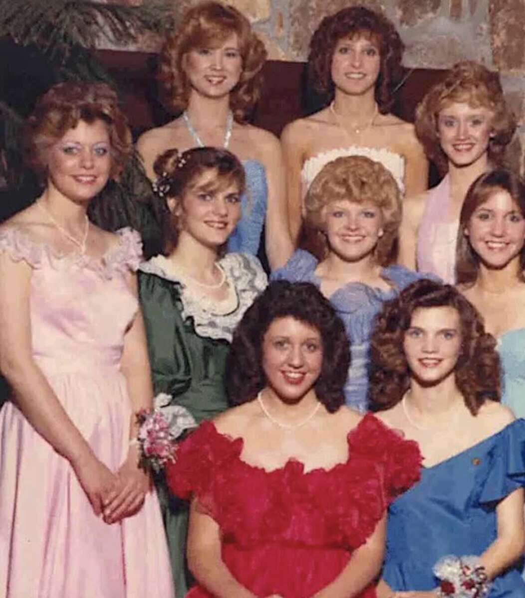 A group of nine young women in colorful, formal 1980s-style dresses stand posed and smiling, some with corsages, against an indoor backdrop with plants.
