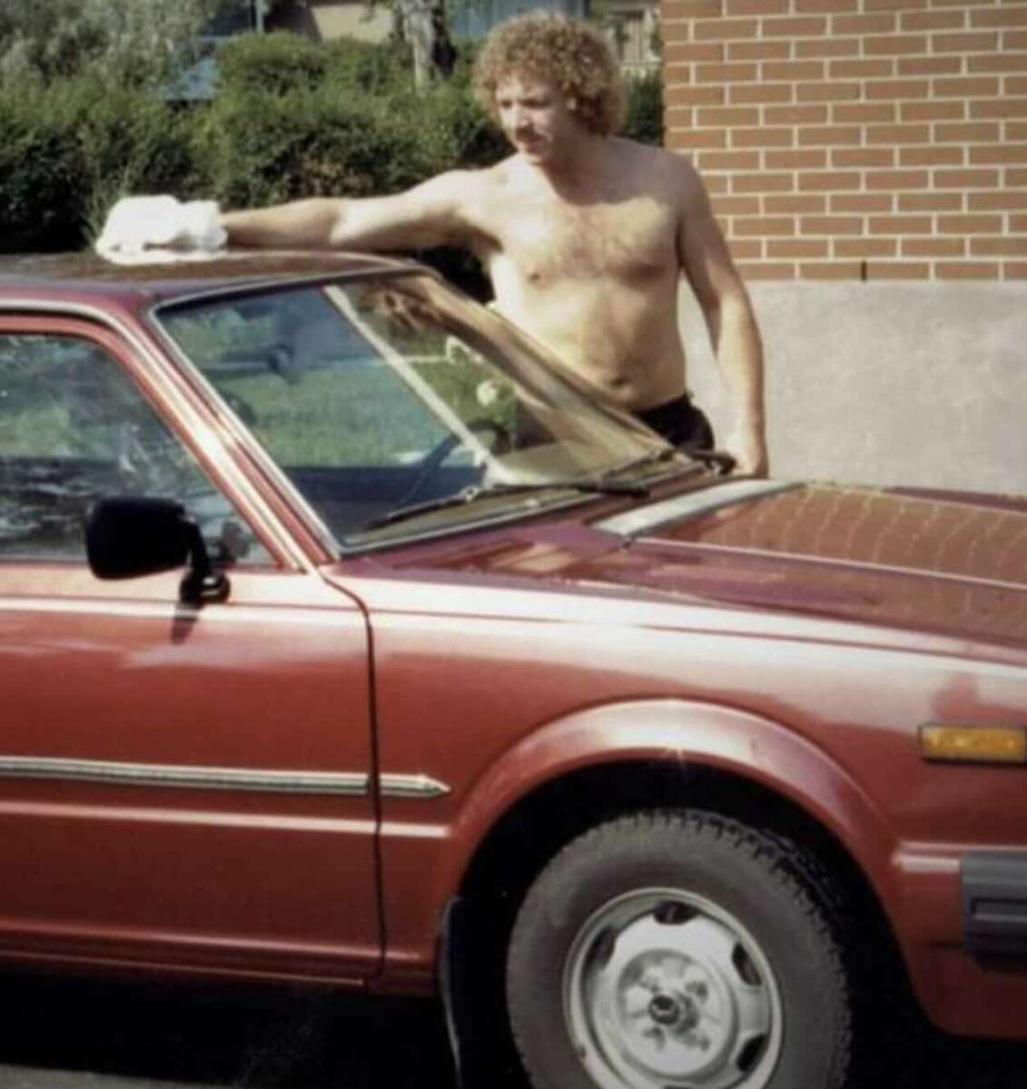 Shirtless man with curly hair cleaning the windshield of a red car with a cloth, standing next to a brick wall on a sunny day.