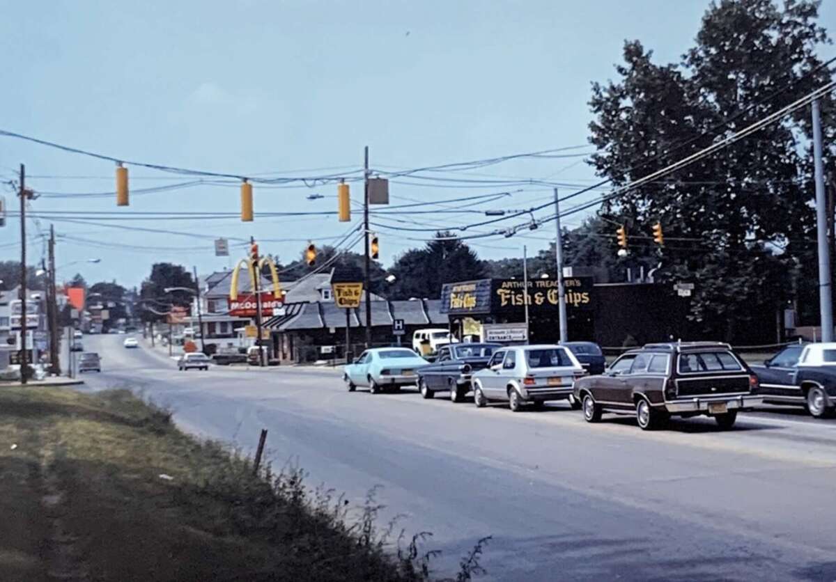 A busy street scene from the 1980s with several cars at a traffic light, fast food restaurants, and a Fish & Chips shop visible along the roadside under a clear sky.