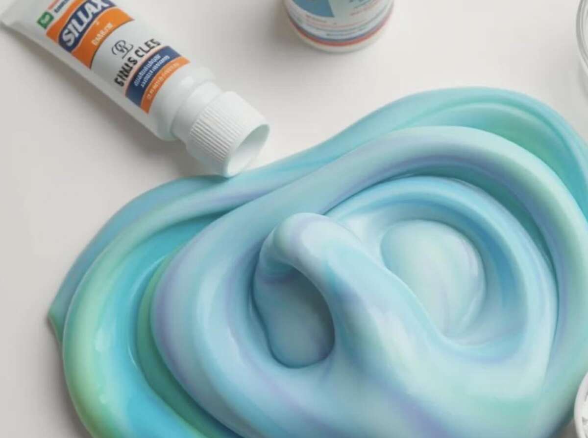 Swirled blue, turquoise, and white clay or putty is spread across a white surface, with a tube of Siluca child-safe clay lying nearby. The clay has a glossy texture and soft, marbled appearance.