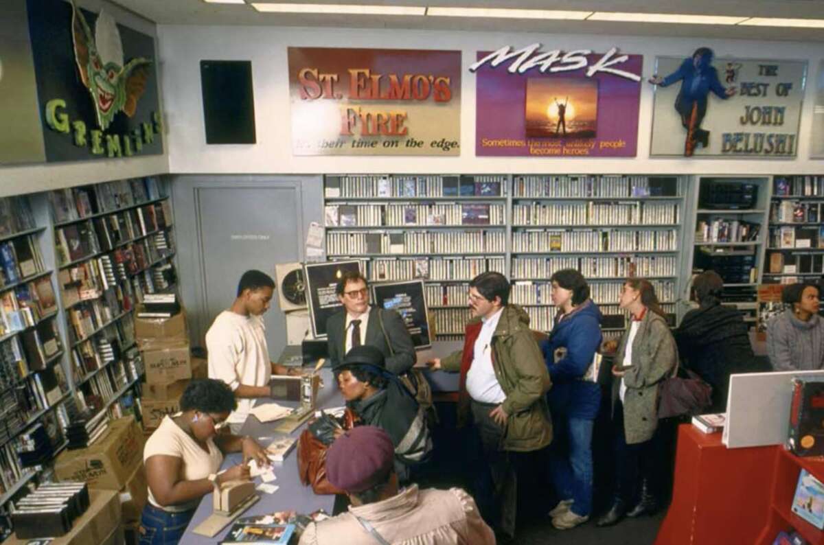 A group of people wait in line at the counter of a video rental store filled with VHS tapes. Posters for "Gremlins," "St. Elmo's Fire," "Mask," and "The Best of John Belushi" are on the walls.