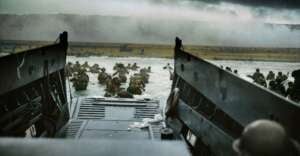 Soldiers wade through ocean water toward a smoky, sandy shore, seen from the back of a landing craft during a wartime beach invasion under a dark, cloudy sky.