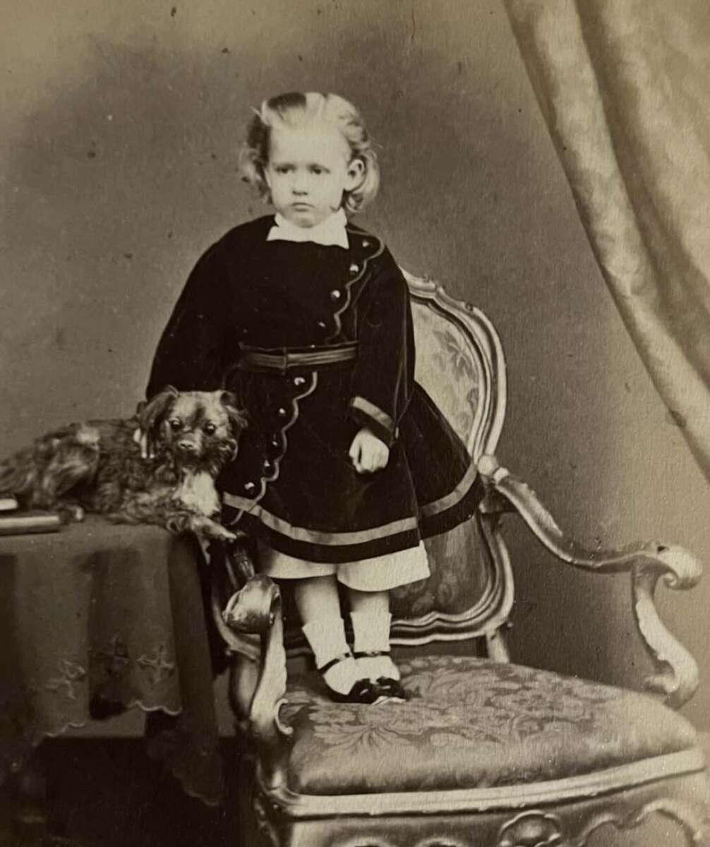 A vintage photograph of a young child in a dark dress standing on an ornate chair, with a small dog resting on a nearby table. A curtained backdrop is visible in the background.