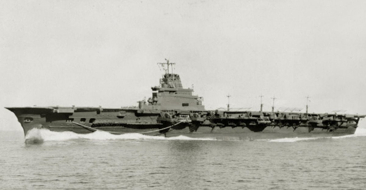 A large aircraft carrier sails at speed across calm waters. Several planes are visible on its deck, and the ship is viewed from the side against a light, featureless sky.