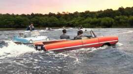 Several classic cars, including a red convertible, drive through water like boats, with people inside. Trees and a sunset sky are in the background. The cars create waves as they travel across the water.