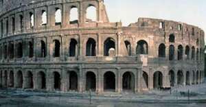 The image shows the ancient Roman Colosseum, a large, partially ruined amphitheater with multiple arches, weathered stone walls, and an open-air structure.