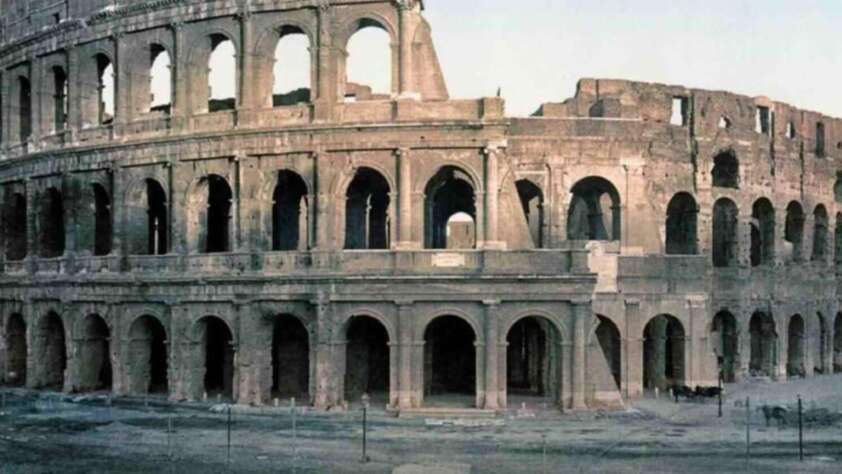 The image shows the ancient Roman Colosseum, a large, partially ruined amphitheater with multiple arches, weathered stone walls, and an open-air structure.