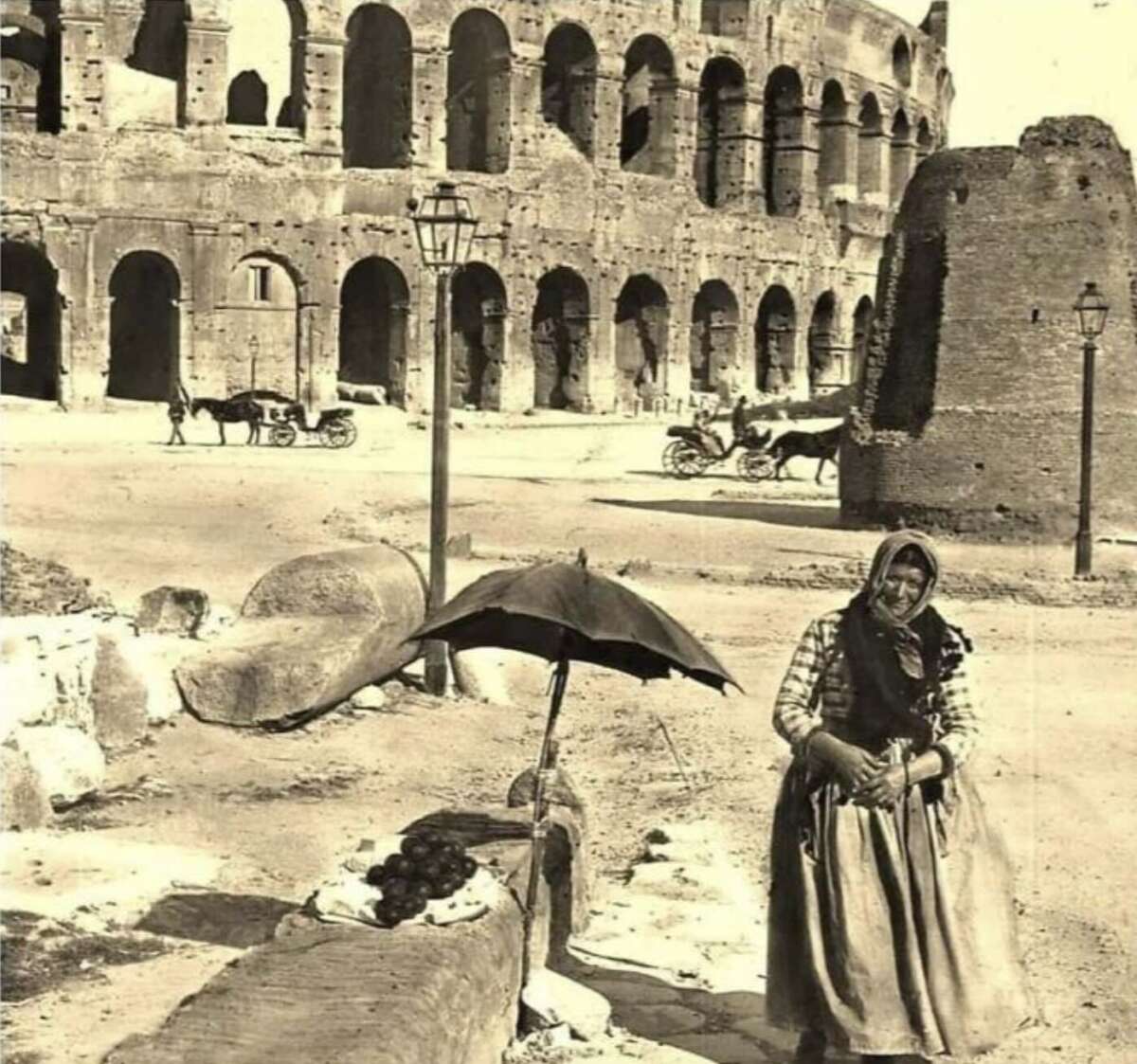 A woman in traditional clothing stands near an umbrella and stone ruins in front of the Colosseum in Rome, with horse-drawn carriages in the background. The photo is in black and white.