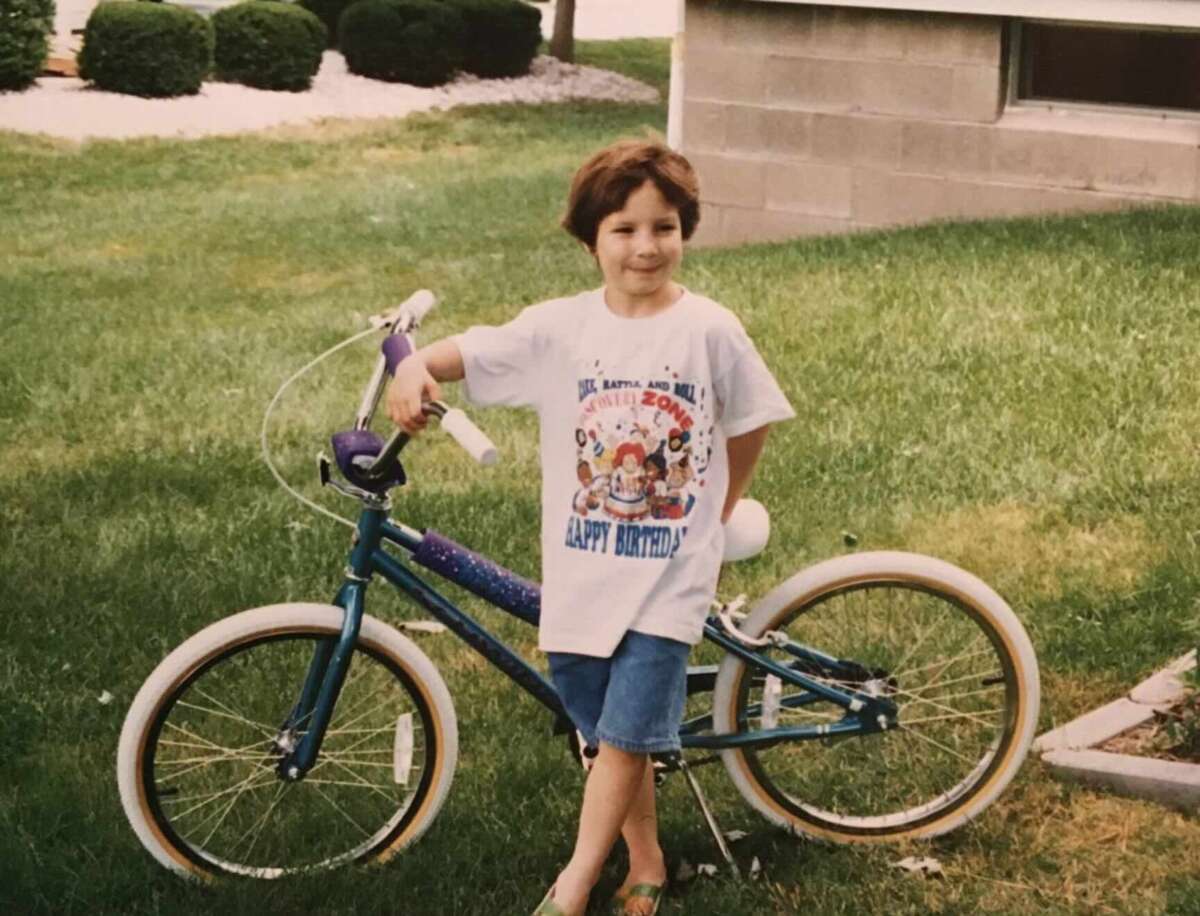 A young child stands outdoors on grass, smiling and posing with one hand on a blue bicycle. The child wears a birthday-themed T-shirt and denim shorts, with trees and a building visible in the background.