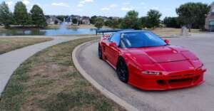 A bright red sports car with black wheels is parked on a curved street near a pond, with trees, grass, and houses visible in the background under a partly cloudy sky.