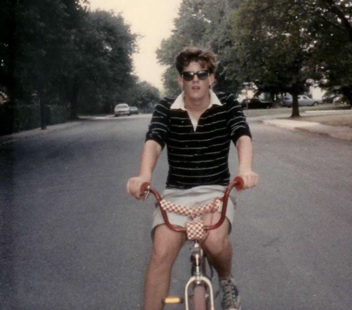 A person wearing sunglasses, a black and white striped shirt, and shorts rides a bicycle down a quiet suburban street lined with trees and parked cars.