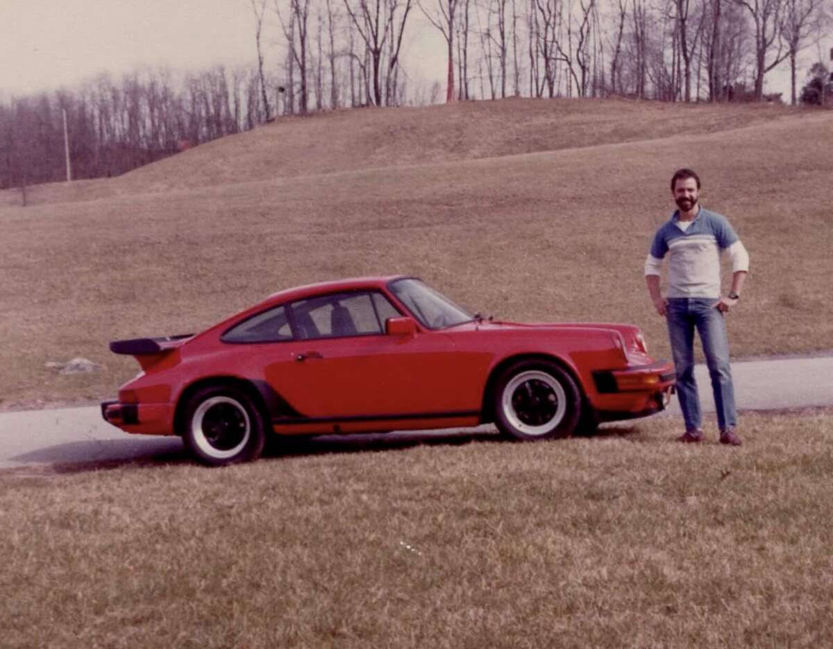 A man with a beard stands smiling next to a red classic sports car parked on grass, with leafless trees and a sloping hill in the background on an overcast day.