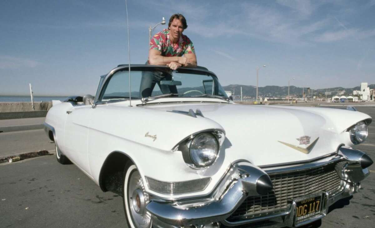 A man in a colorful shirt leans on the windshield of a classic white Cadillac convertible parked by the seaside on a sunny day, with buildings and mountains in the background.