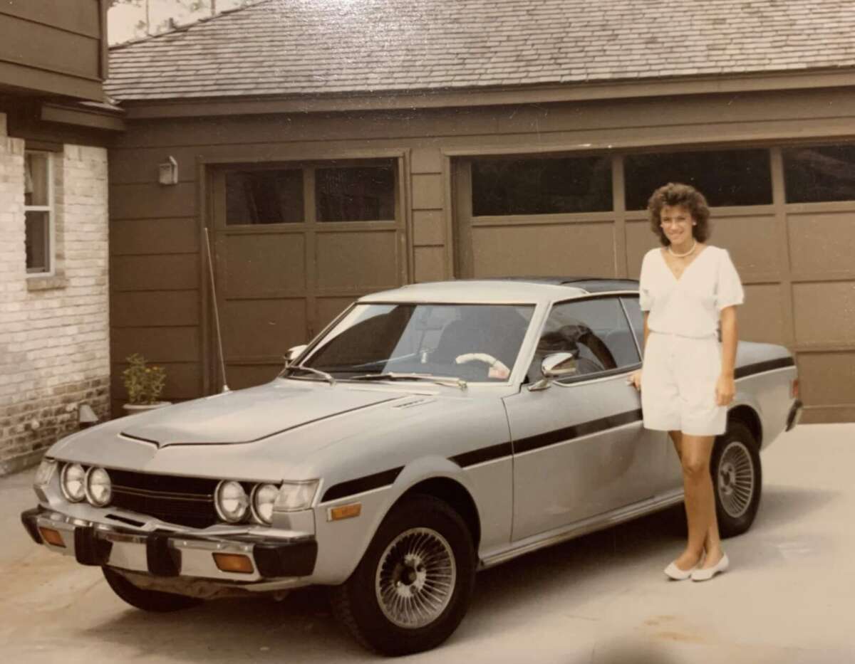 A woman in a white outfit stands and smiles next to a vintage silver car parked in a driveway in front of a brown garage.