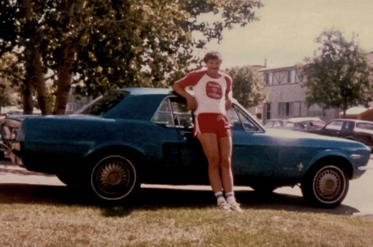 A person wearing a red and white athletic outfit stands in front of a blue vintage car parked on grass, with trees and houses visible in the background on a sunny day.