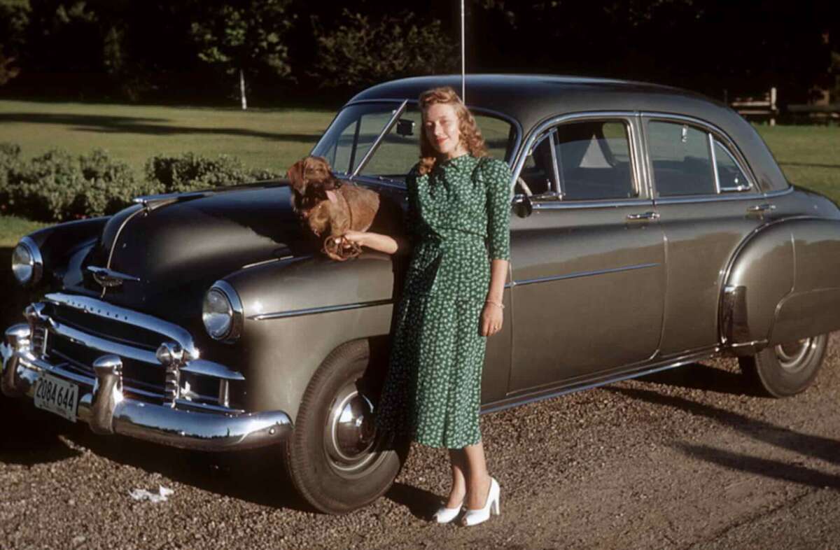 A woman in a green patterned dress and white shoes leans against a vintage gray car, smiling, with a small brown dog sitting on the car’s hood. Green grass and trees are in the background.