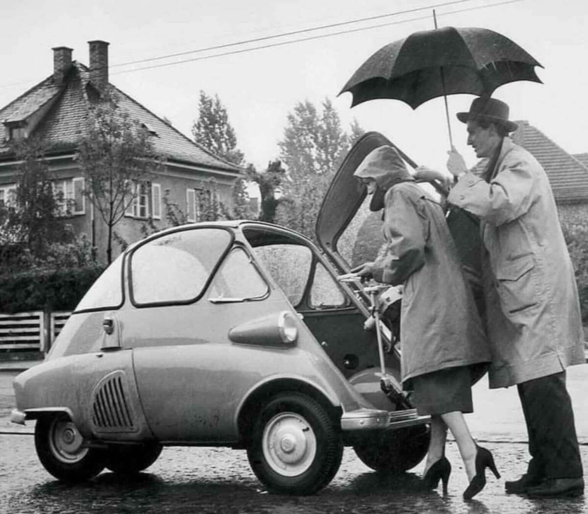 A man holds an umbrella over a woman as she puts something in the front trunk of a small three-wheeled microcar on a rainy day; both are wearing trench coats. A house and wet street are in the background.