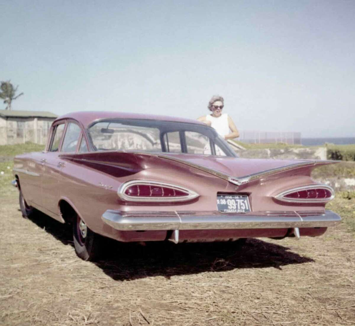 A woman wearing sunglasses stands next to a pink 1959 Chevrolet car with prominent tailfins, parked on dry grass near a building and the ocean under a clear sky.