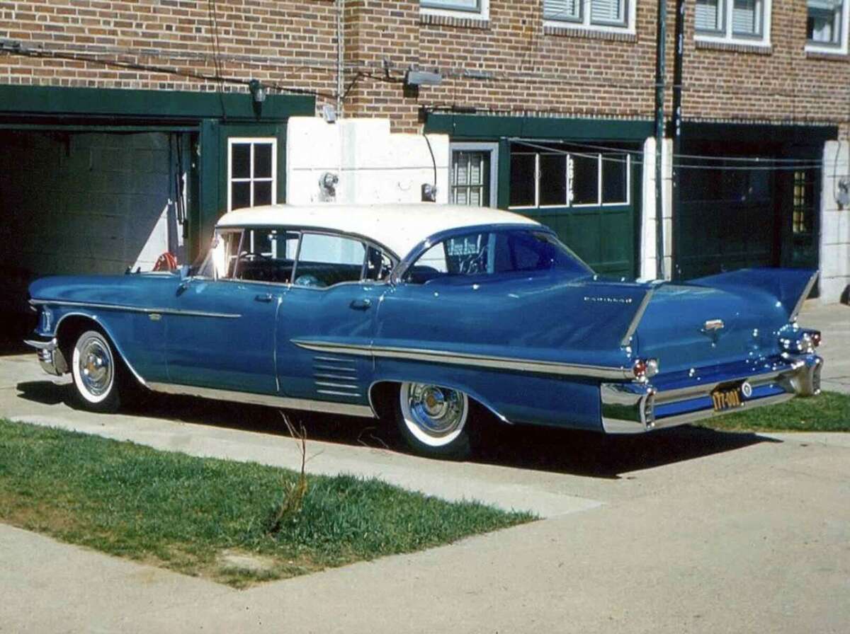 A classic blue and white vintage car with fins is parked in a driveway in front of a brick building with green garage doors and windows.