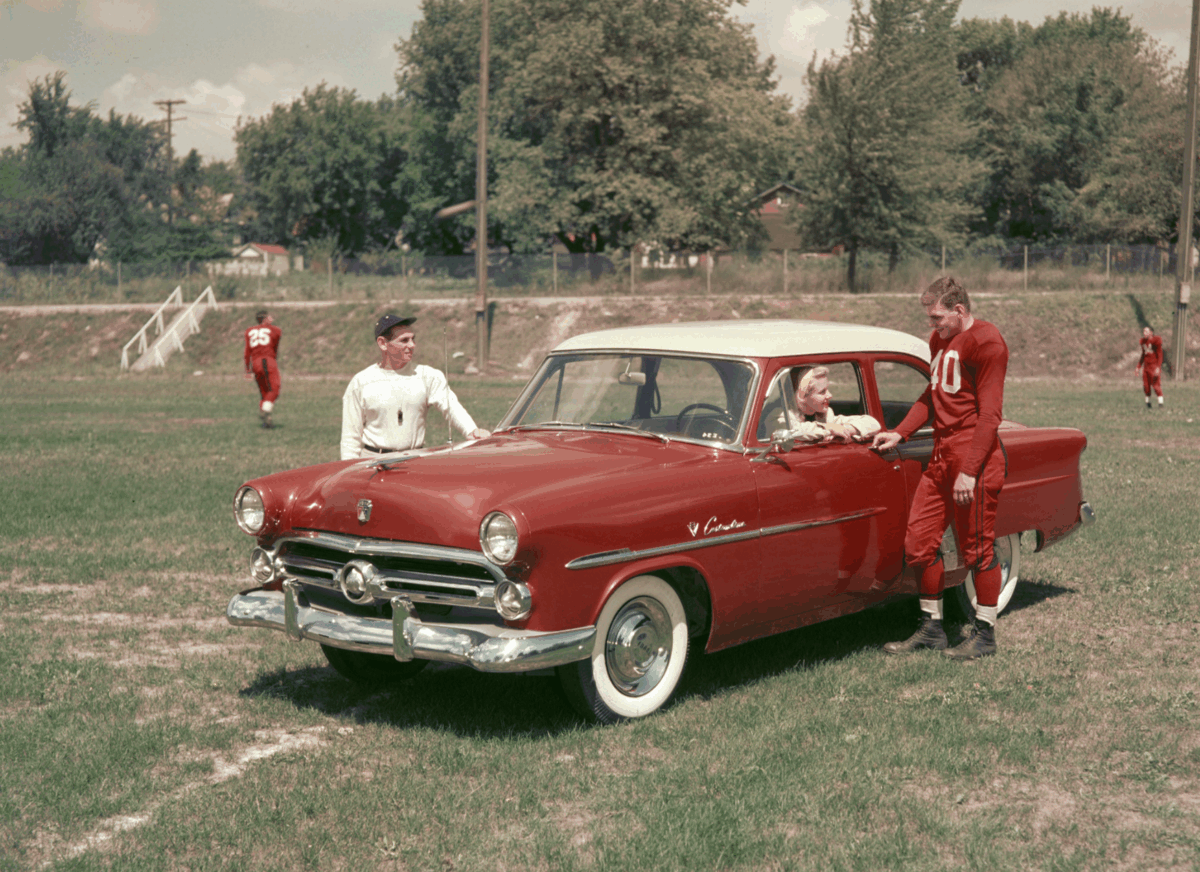 Three people in football uniforms stand by a vintage red car on a grassy field, with one sitting inside the car and two standing outside. More football players can be seen practicing in the background.