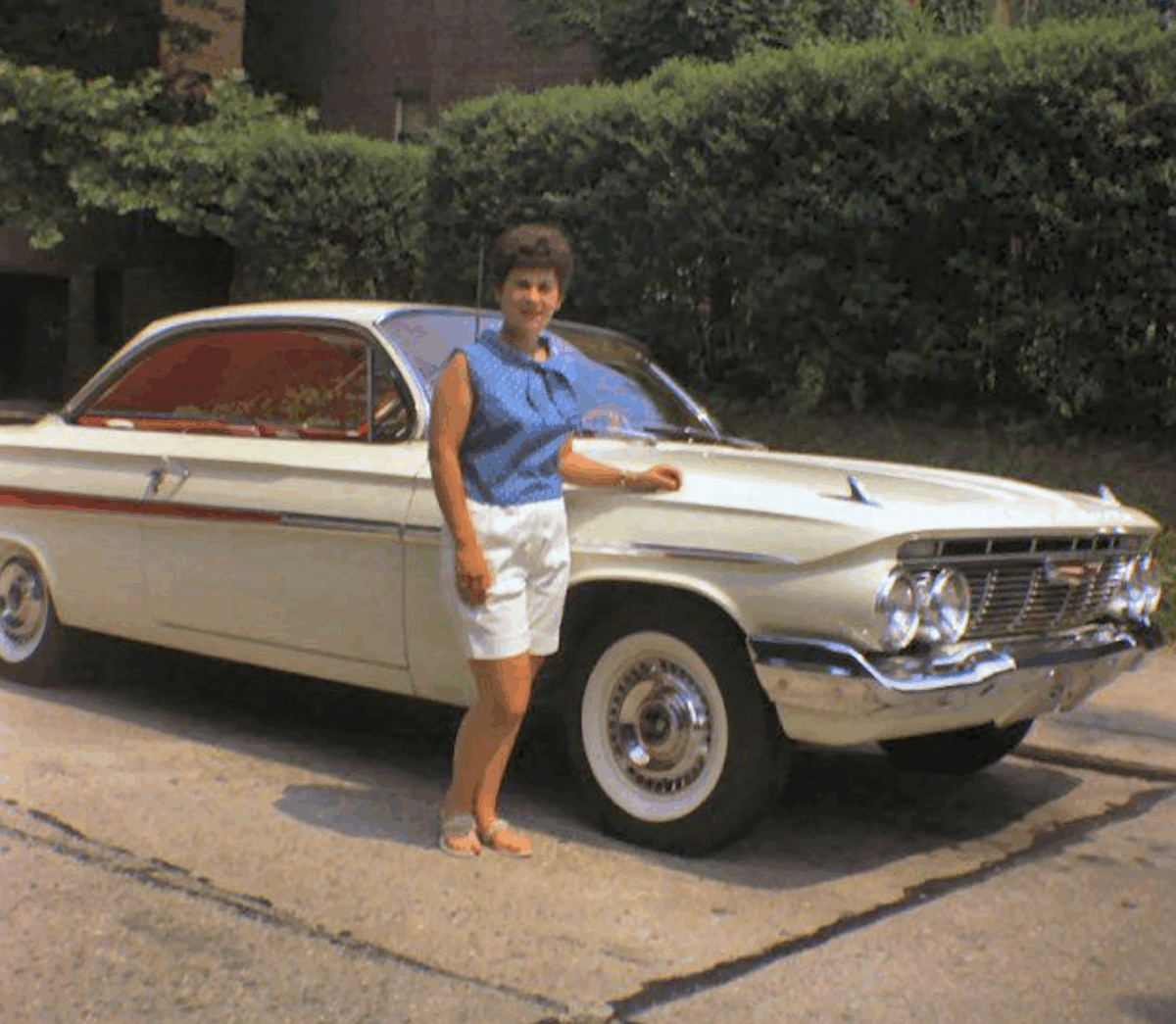 A woman in a blue sleeveless top and white shorts stands next to a vintage white car with red accents, parked on a sunny street with greenery in the background.
