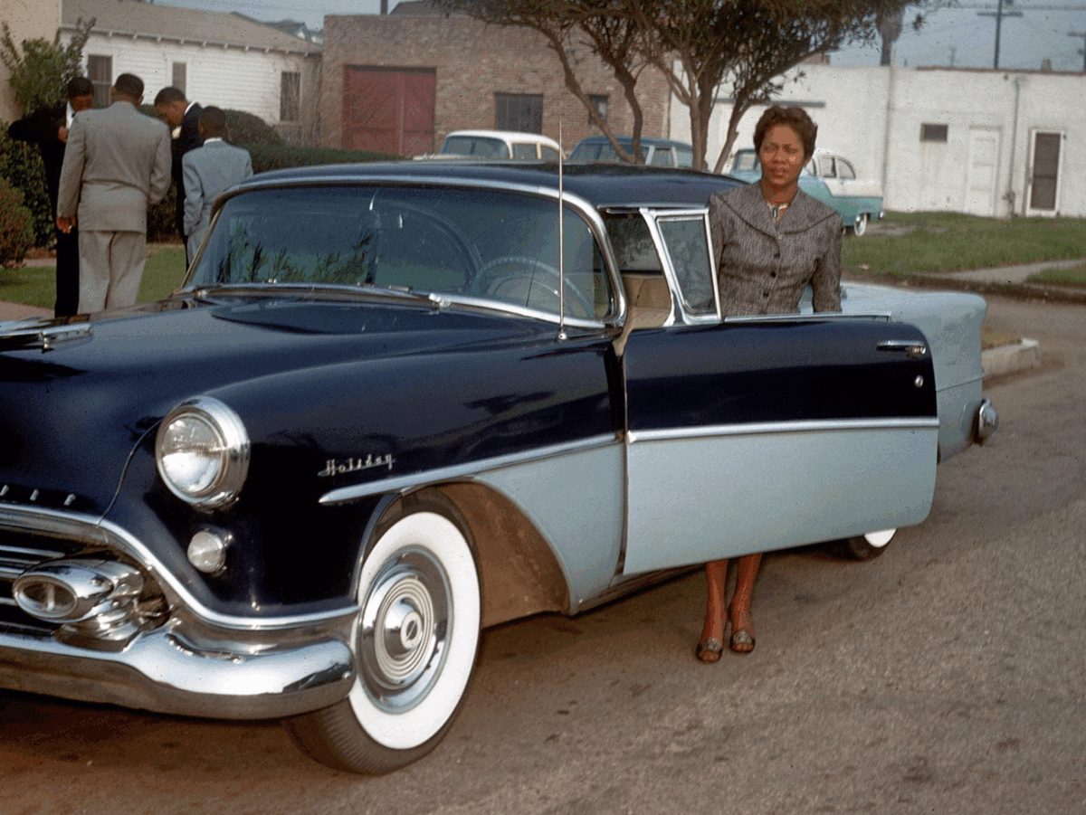 A woman in a gray suit stands by the open door of a vintage blue and white car parked on a street, with several people and more cars visible in the background.