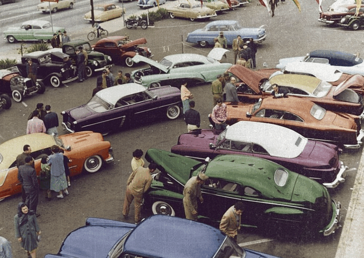 A vintage car show with people walking around and admiring colorful classic cars parked closely together in an outdoor parking lot. The cars are in shades of green, purple, orange, red, blue, and cream.