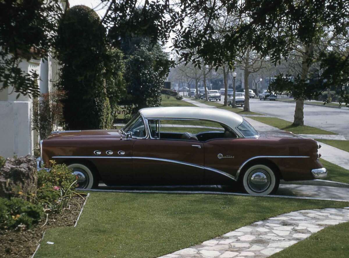 A vintage maroon and white Buick car is parked on a residential street next to manicured grass and a stone walkway, with trees lining the road in the background.
