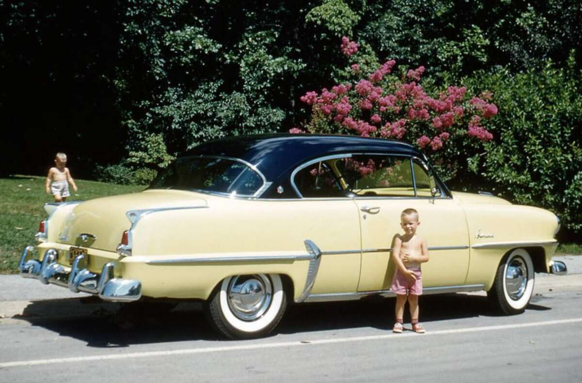 A vintage yellow and black car is parked by the curb. A shirtless young boy in pink shorts stands in front of the car, while another child is in the background. Green trees and flowering bushes are visible behind them.