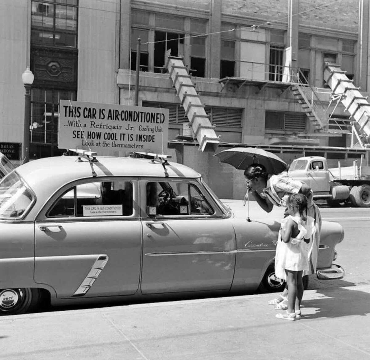 A woman leans into a vintage car while holding an umbrella, as a young girl stands beside her. A sign on top of the car reads, “This car is air conditioned.” A city building is visible in the background.