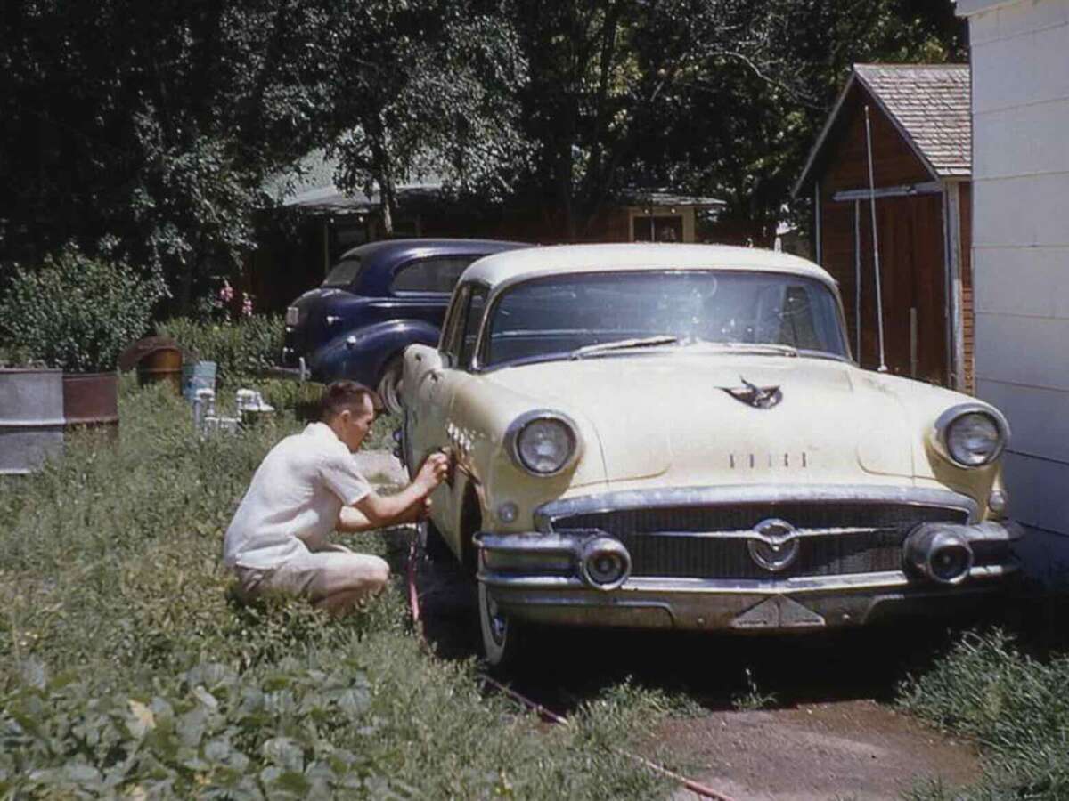 A man in shorts and a light shirt crouches beside a vintage yellow car, washing or working on its front fender, in a grassy yard with another classic car, trees, and a wooden shed in the background.