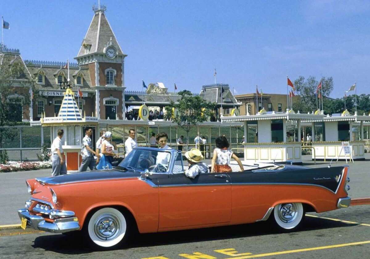 A vintage red and white convertible car is parked in front of Disneyland's entrance. Several people, including a family in summer clothing, are near the car with Disneyland's Main Street train station and ticket booths in the background.