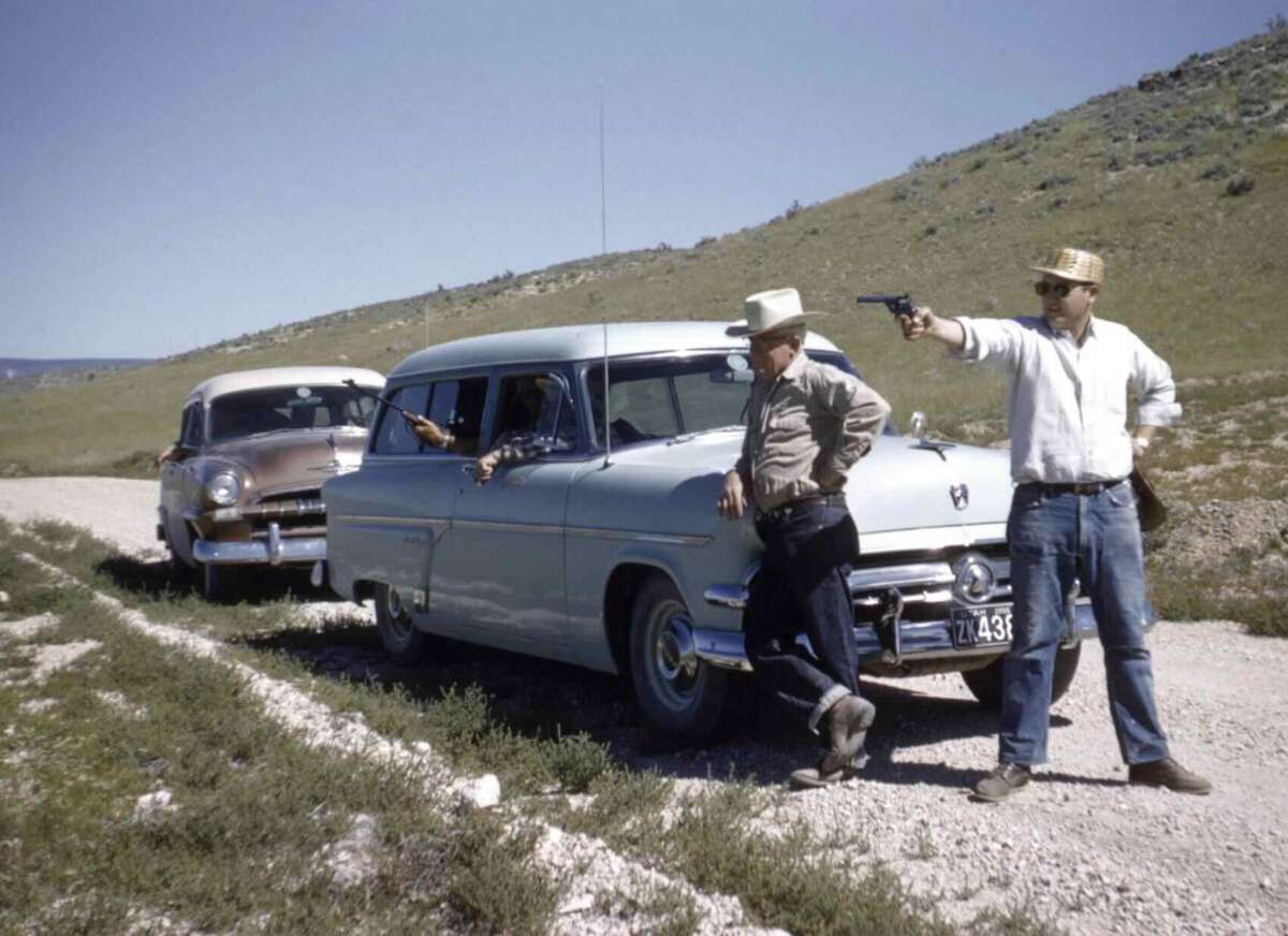 A man in a white shirt and straw hat points a gun at another man leaning against a vintage blue car on a rural dirt road, with a second vintage car and a driver in the background.