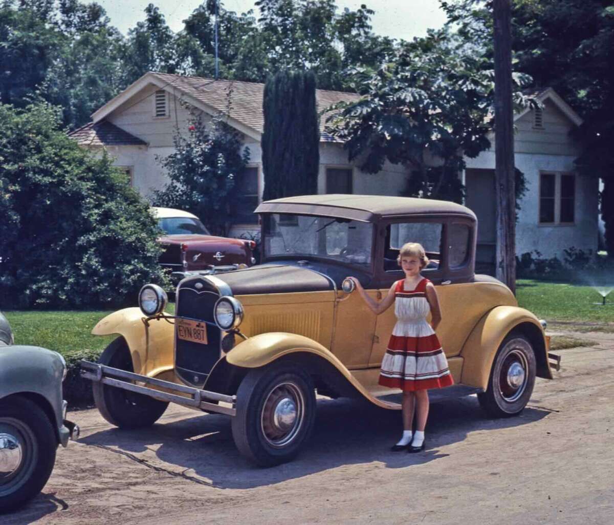 A young girl in a red and white dress stands beside a vintage yellow car parked on a residential street, with trees and houses visible in the background on a sunny day.