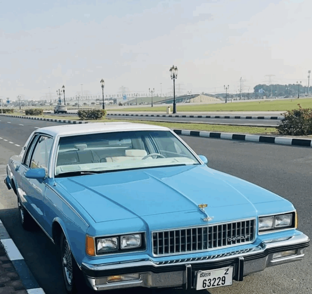 A bright blue vintage car is parked on the side of a wide, empty road with street lamps and greenery in the background under a clear sky.