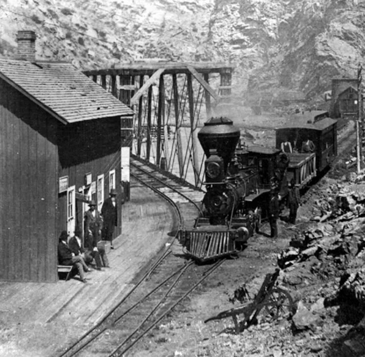 Black-and-white photo of a steam locomotive approaching a wooden train station beside rocky terrain, with several people sitting and standing nearby and a metal truss bridge in the background.