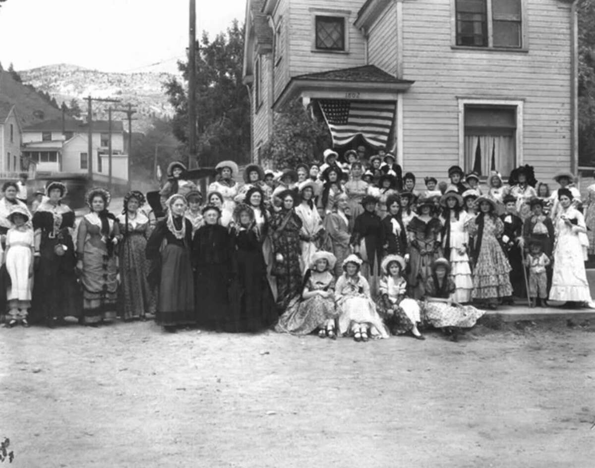 A large group of women in vintage dresses and hats pose in front of a wooden house with an American flag; trees, mountains, and other houses are visible in the background.