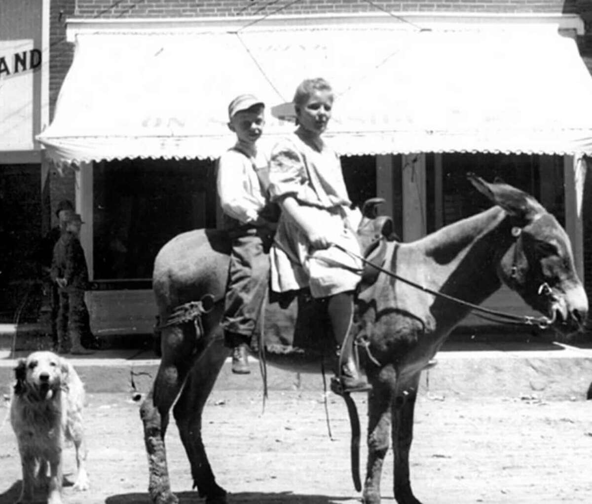 Two children sit on a donkey in front of a storefront, while a dog stands nearby. The children are dressed in old-fashioned clothes, and the scene appears to be from an earlier era.