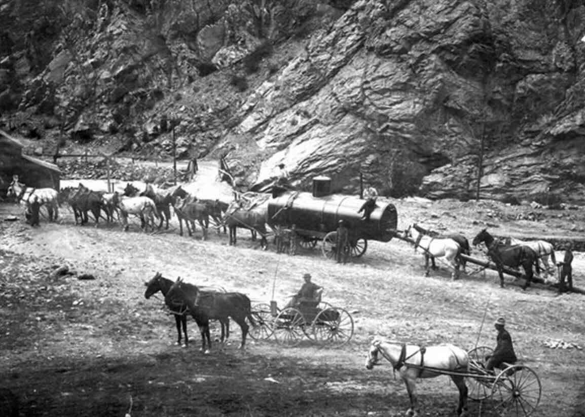 A historic black-and-white photo showing several horse-drawn carts and wagons traveling along a rugged dirt road beside rocky cliffs. Men sit in the wagons, and a large tank is being pulled by a team of horses.