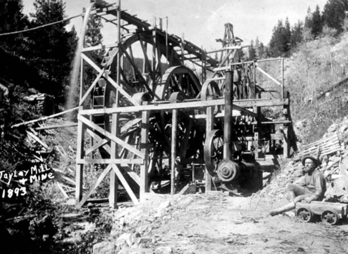 Black and white photo of a large wooden mining structure with wheels and machinery, labeled “Taylor Mill & Mine 1893.” A person sits on the ground nearby, surrounded by rocky terrain and trees.