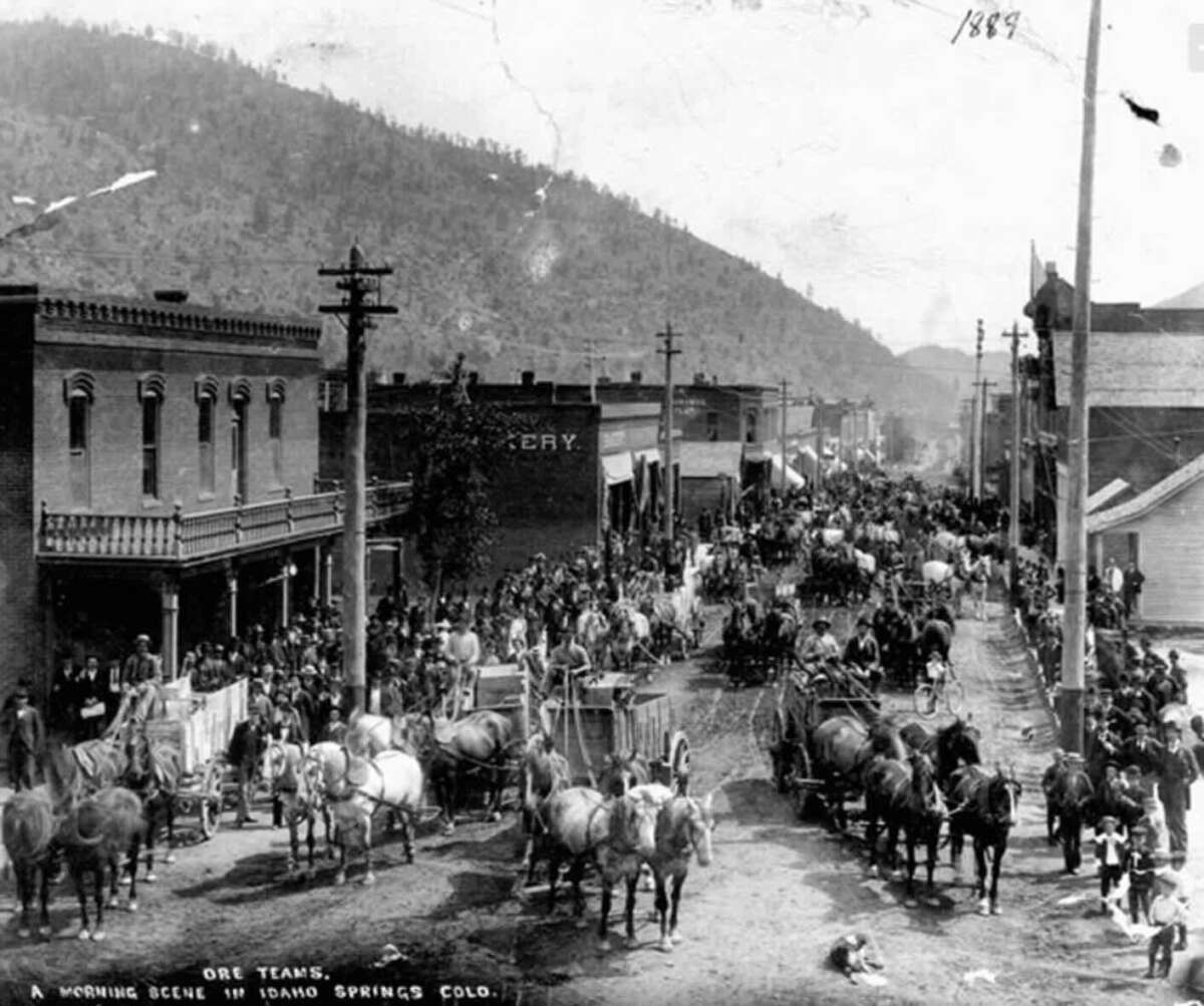 A black-and-white photo from 1889 shows a busy street in Idaho Springs, Colorado, filled with horse-drawn wagons and people lined up along both sides of the street. Mountains rise in the background.