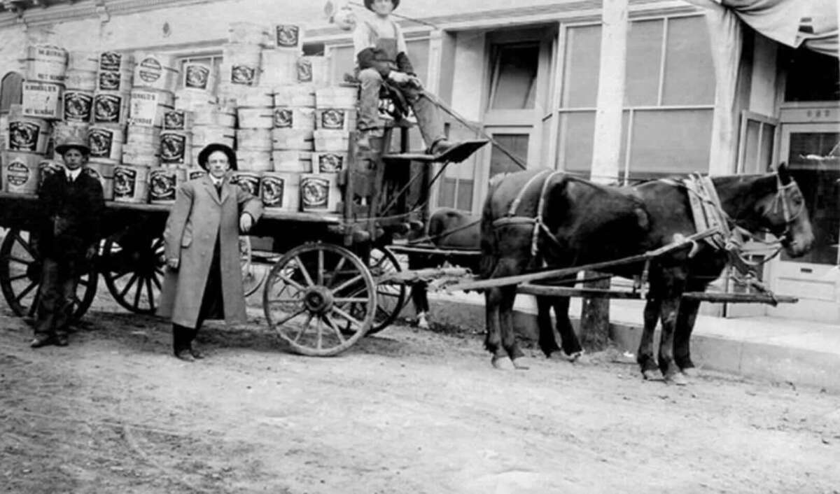 Black and white photo of three people and a child with a horse-drawn wagon loaded with barrels, posed in front of a building. Two stand beside the wagon; one child sits on top.