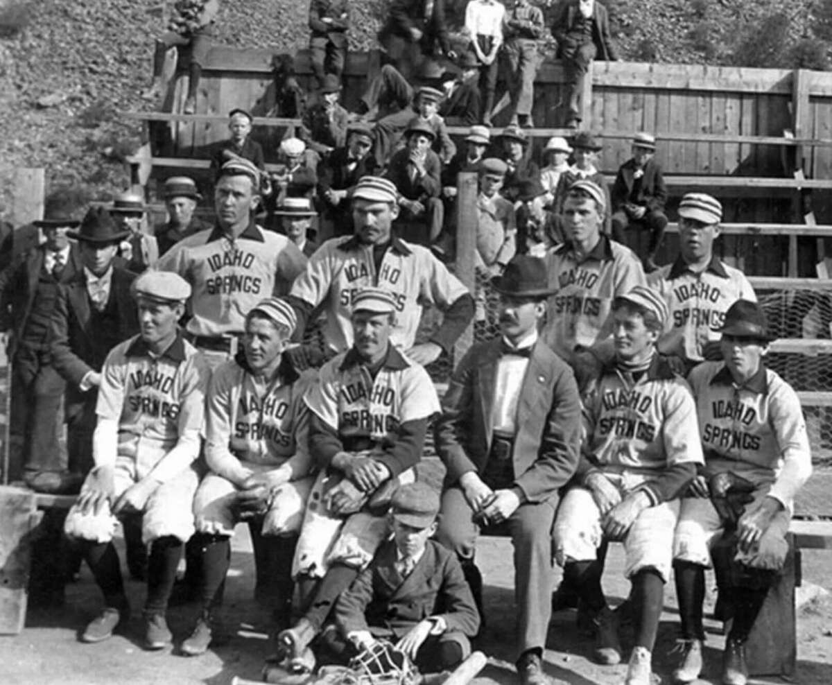 Black and white photo of a vintage baseball team in “Idaho Springs” uniforms, posing with coaches and a young boy in front of bleachers filled with spectators.