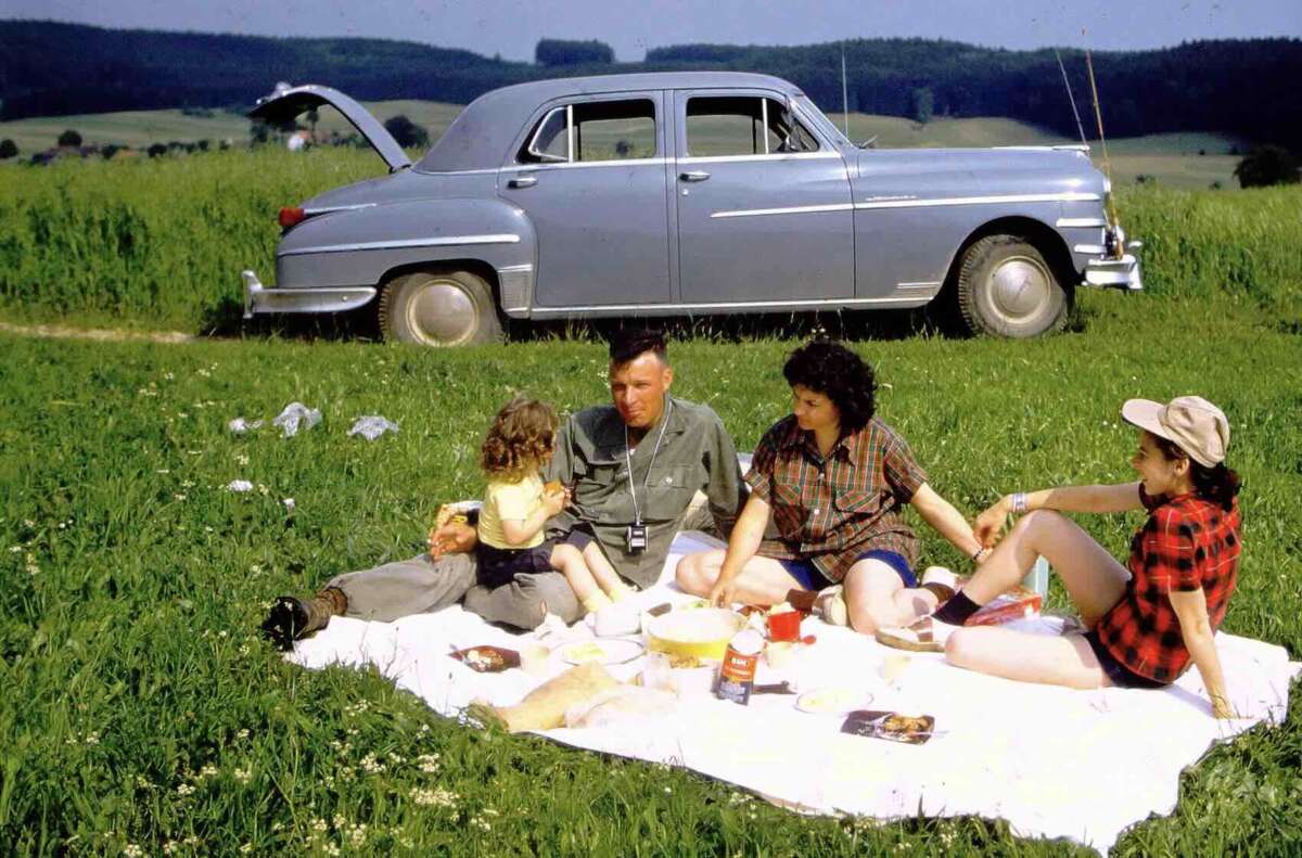 A family of four enjoys a picnic on a white blanket in a grassy field, with food and drinks spread out. A vintage car with its trunk open is parked nearby; rolling hills and trees are in the background.