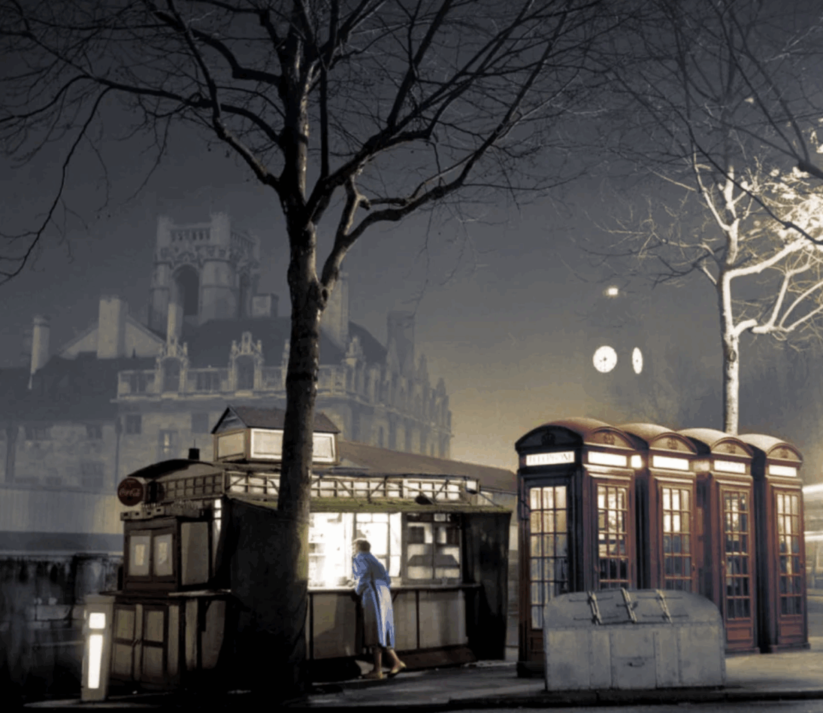A person in a blue coat stands at a small kiosk at night, next to a row of classic red British phone booths. Bare tree branches and a foggy building are visible in the background.