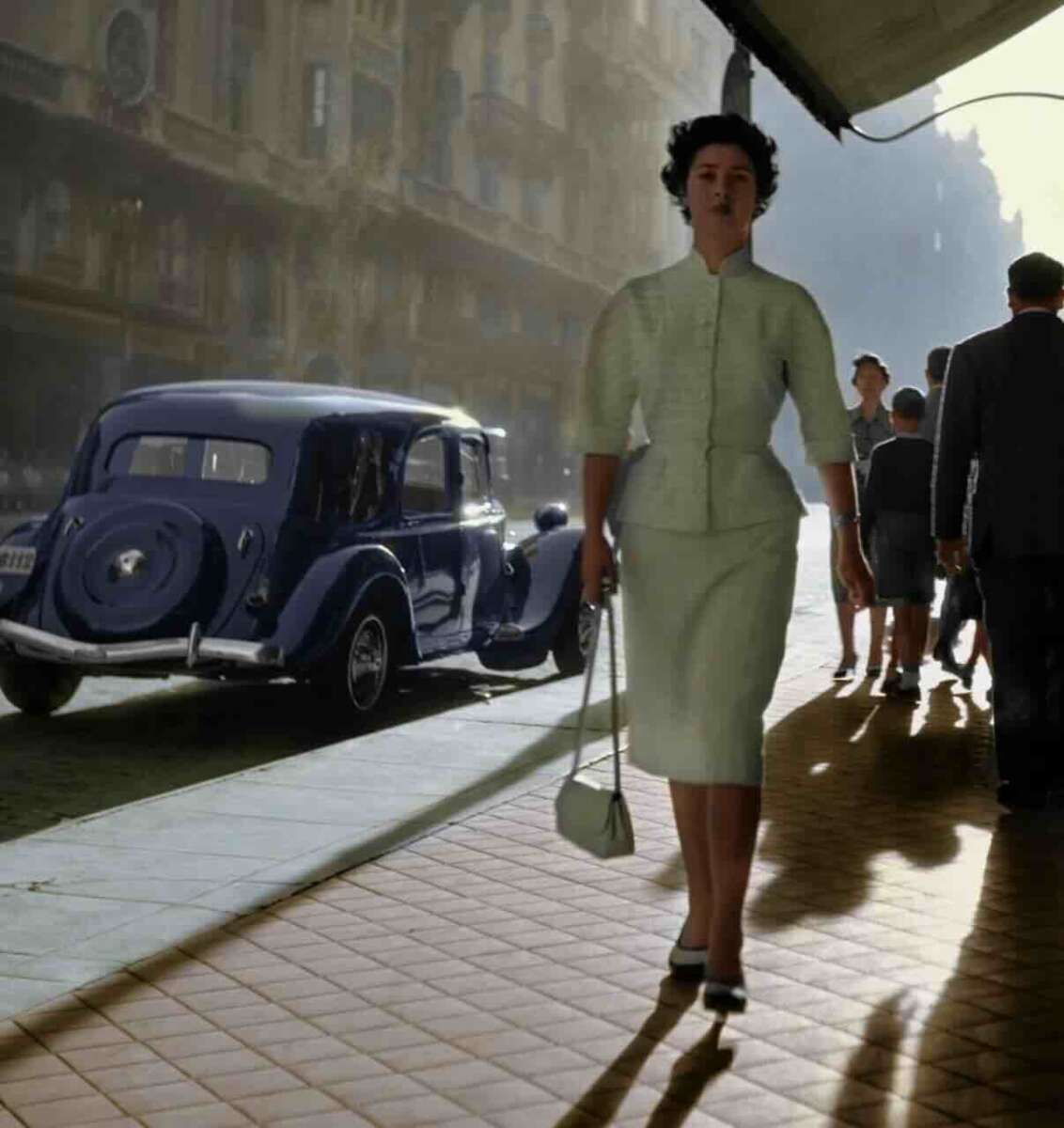 A woman in a light green dress and carrying a purse walks on a sunlit sidewalk beside a vintage blue car, with several people and historic buildings in the background.