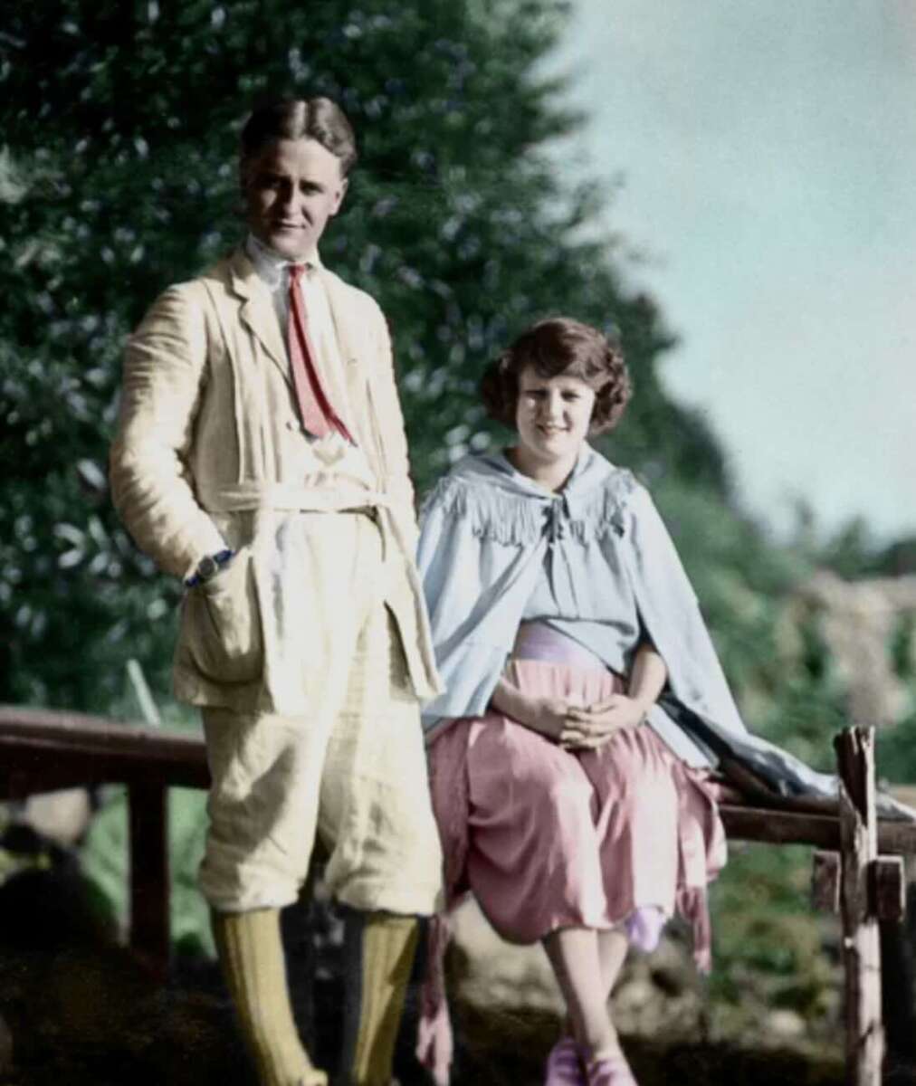 A young man in a light-colored suit stands next to a smiling young woman in a pink skirt and light blue cape, both outdoors near a wooden fence with trees and greenery in the background.