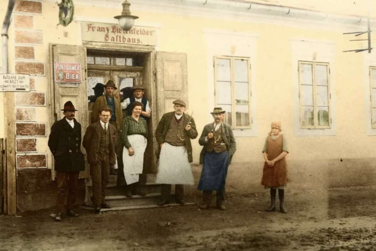 A group of men and one girl stand outside a yellow building with the sign "Franz Blümlfelder Gasthaus." Some wear hats and aprons; the girl stands apart from the group. The scene appears old-fashioned or historical.