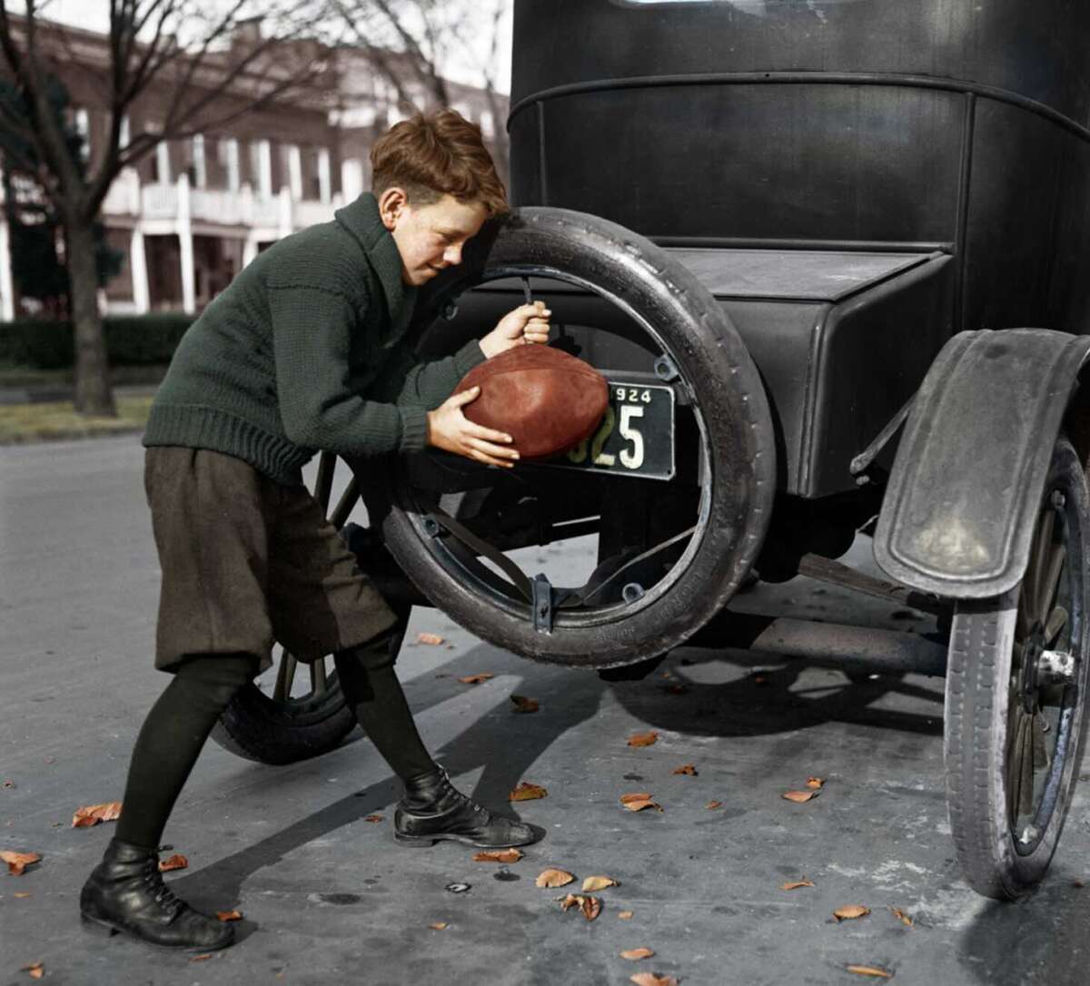 A boy in vintage clothing inflates a football using a car tire at the back of an old-fashioned automobile parked on a leaf-strewn street.